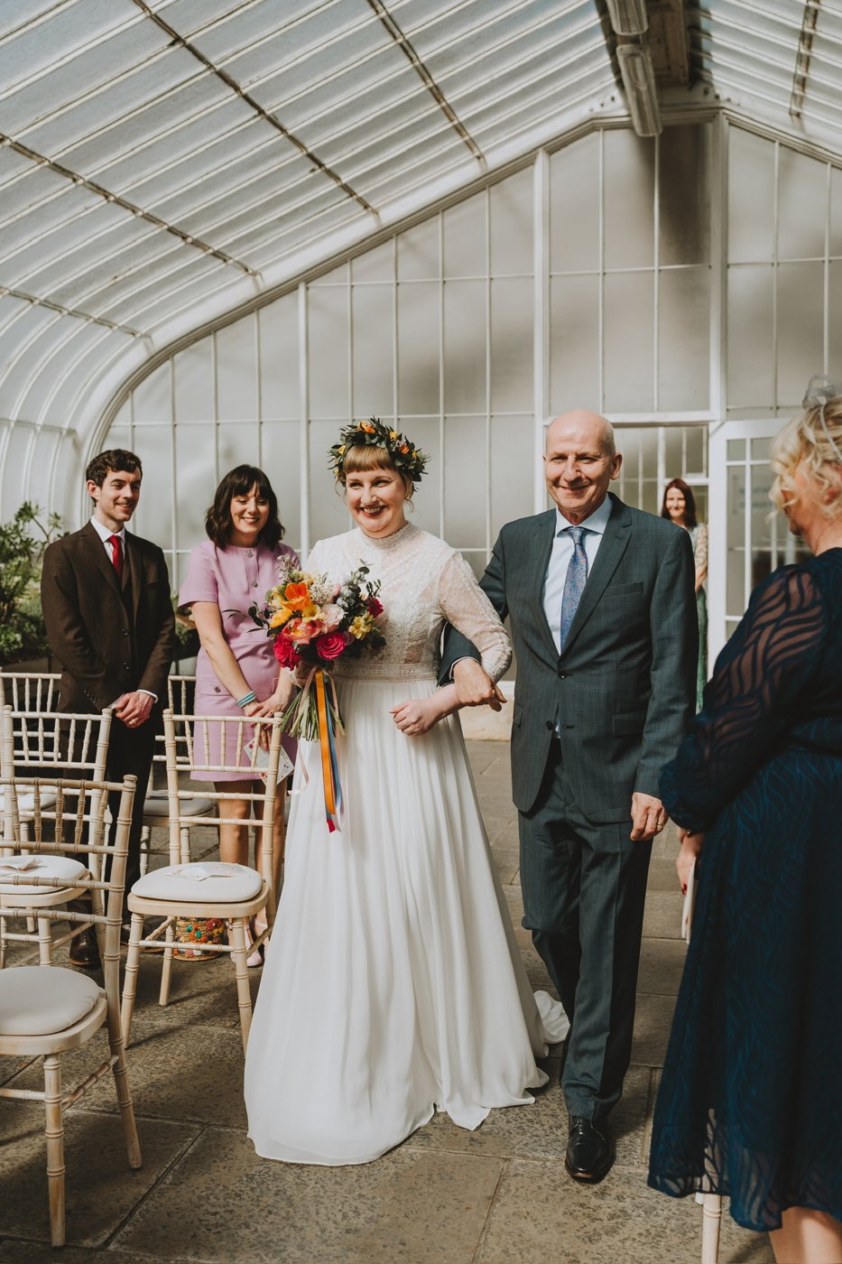 Laura walking down the aisle of the Kibble Palace on her father's arm, wearing a flower crown, guests smiling among the glasshouse ironwork